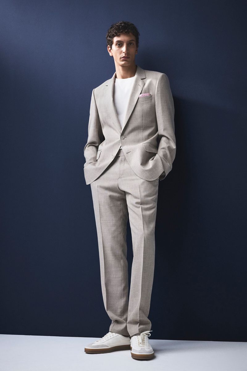 A slender adult male poses in a light grey suit with a white tee shirt and white sneakers against a navy backdrop in a full shot.