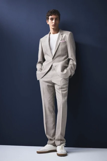 A slender adult male poses in a light grey suit with a white tee shirt and white sneakers against a navy backdrop in a full shot.