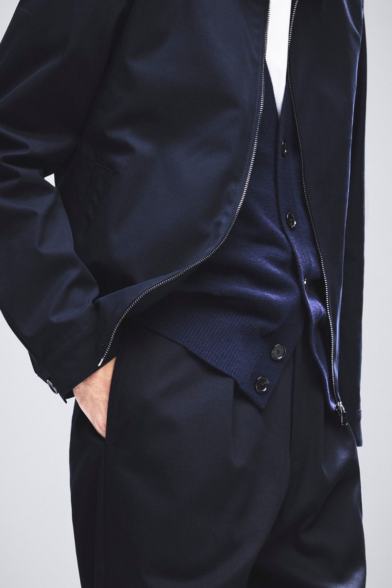 Close-up of an adult wearing a navy jacket, cardigan, and pants against a light background. Fashion and style are highlighted.