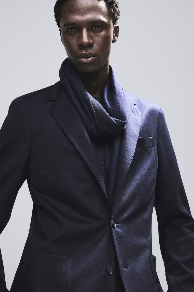 Dark-skinned adult male wearing a navy blue blazer and scarf, posing in a studio with soft, neutral lighting, fashion portrait, menswear.