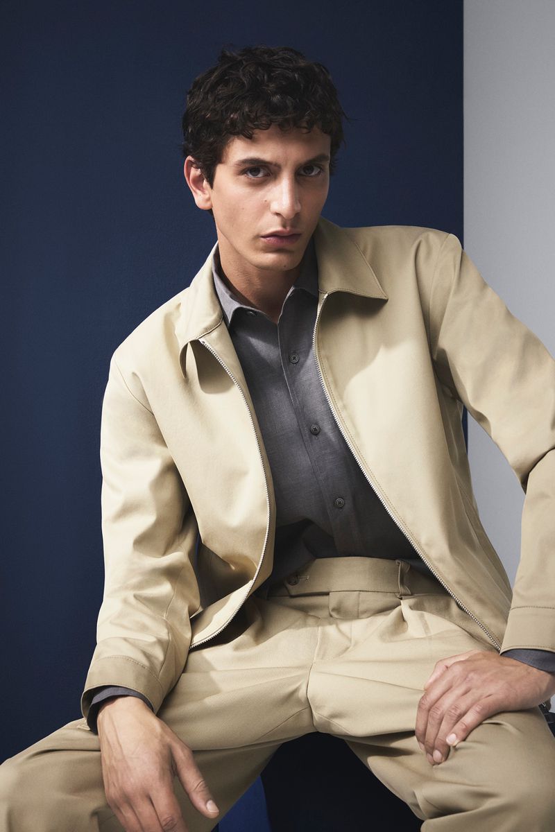 A young adult male poses in a tan jacket and pants, with a grey shirt, against a backdrop of navy and light grey. Fashion headshot.