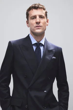 Model George Barnett in a tailored navy double-breasted suit for BOSS, photographed by Marco Cella against a plain light grey studio backdrop.