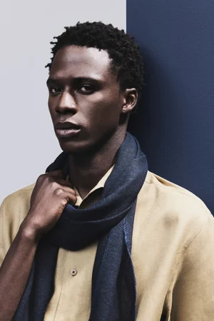 Black male model in a beige button-up shirt and dark navy scarf against a split blue and gray backdrop.