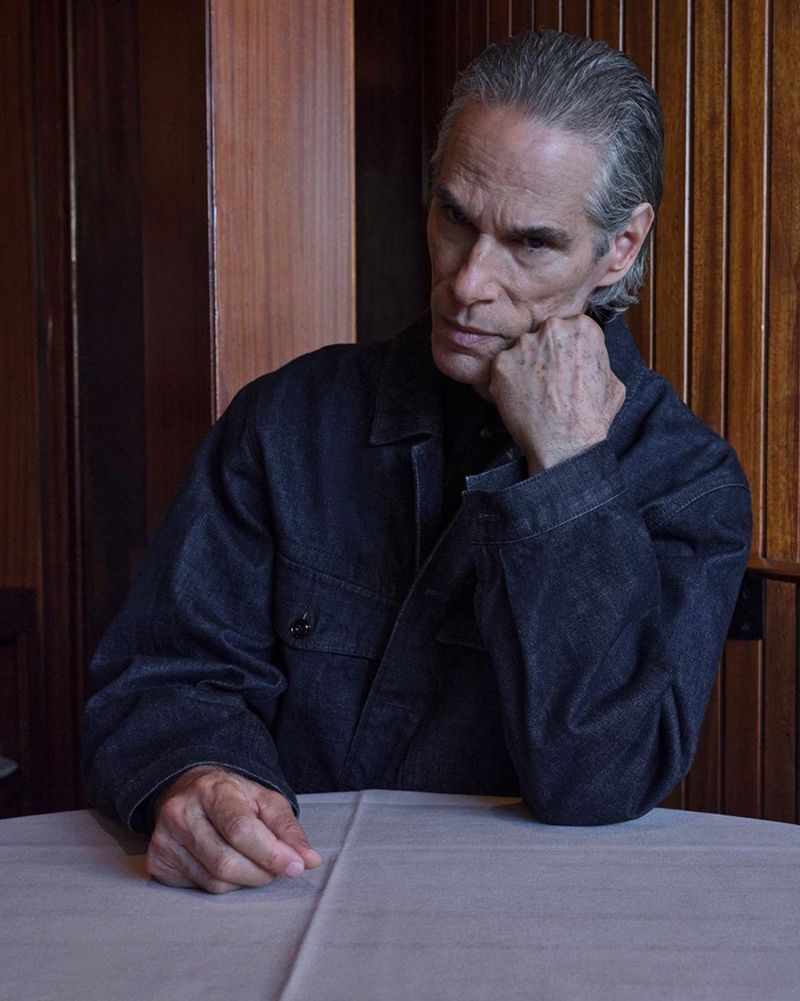 Portrait of a mature man in a dark denim jacket sitting at a table with a white tablecloth, hand to face, contemplative expression.