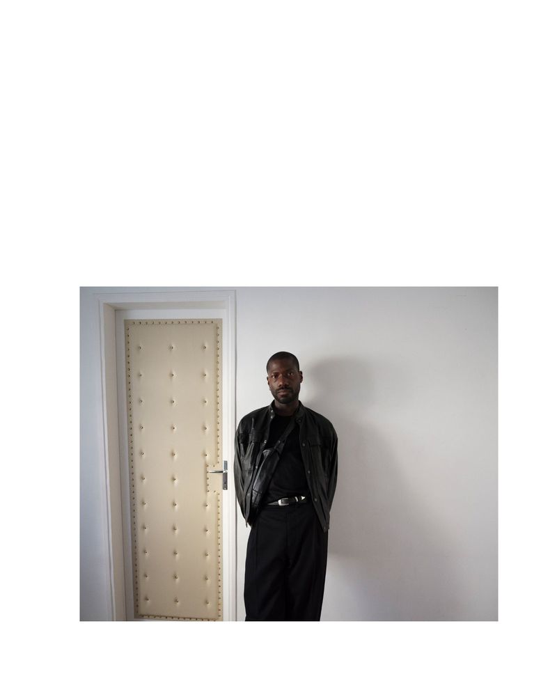 Adult male in a black leather jacket poses indoors in front of a cream-colored door against a white wall, looking directly at the camera.