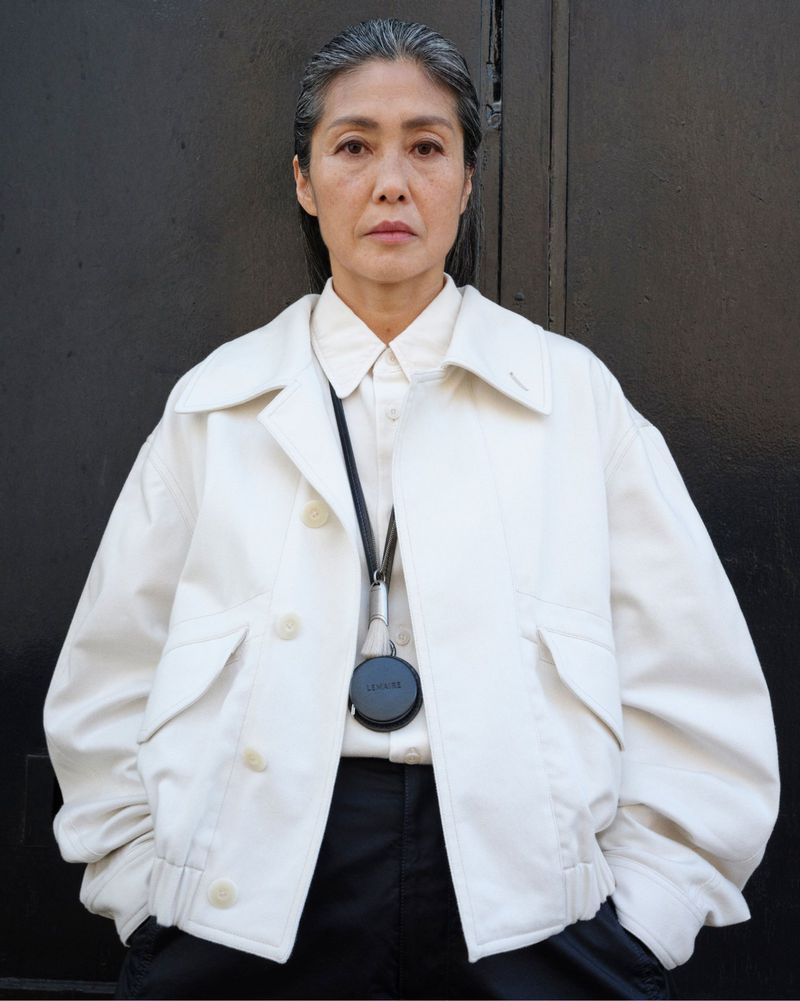 Mature Asian woman in a white jacket and black pants stands against a dark background, captured in a medium portrait shot.