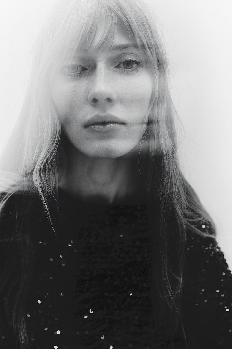 Close-up black-and-white portrait of a young adult woman with long blonde hair, wearing a dark top with sequins against a light background.