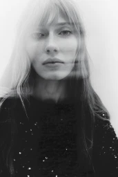 Close-up portrait of a female model with long blonde hair wearing a black sequin top against a plain light background.