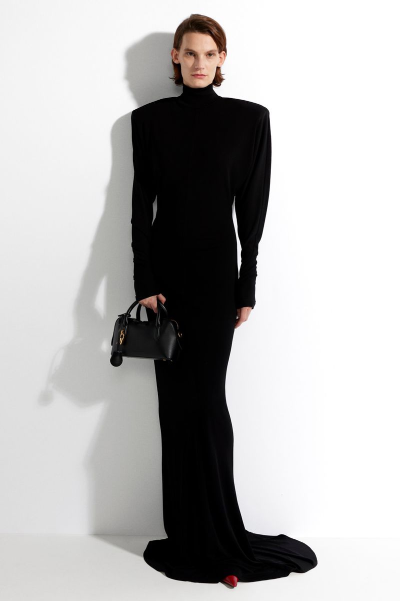 A slender person poses in a long black dress and stylish black handbag against a white background in a fashion-forward studio shot.