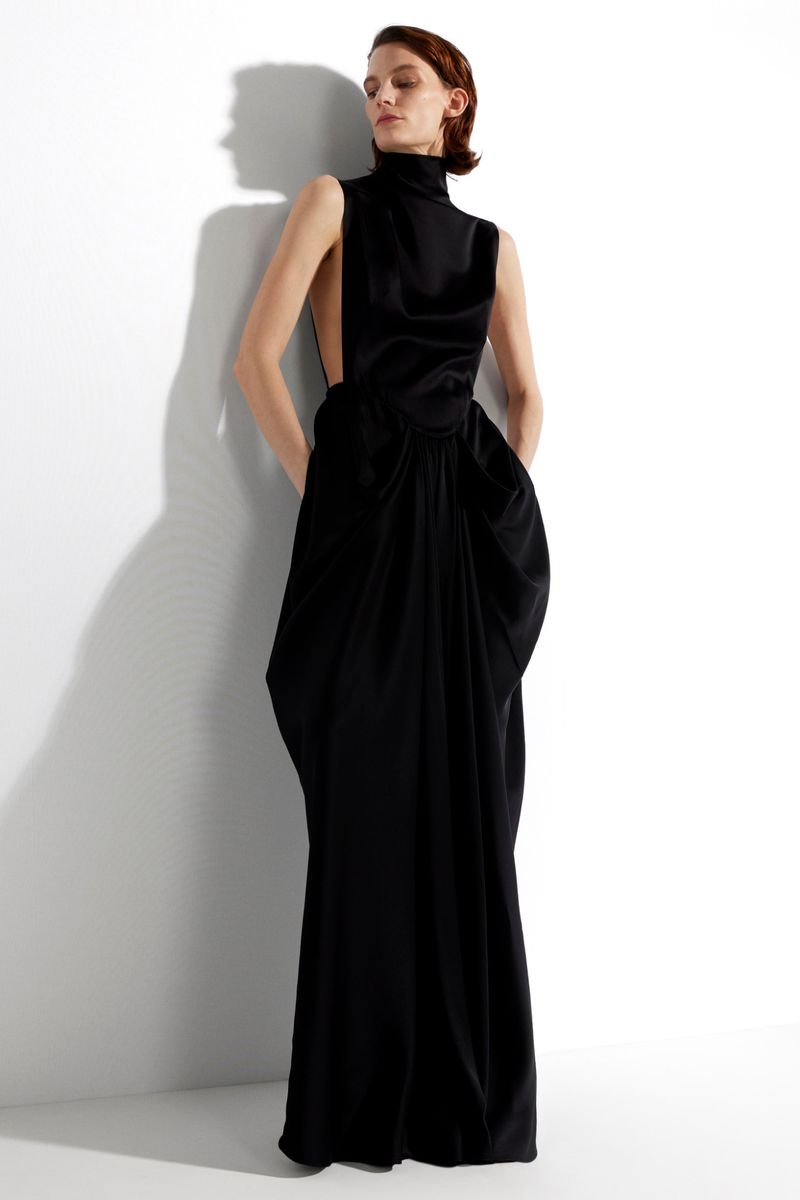 An adult woman poses in a sleeveless black, floor-length gown with a high neckline in front of a light grey studio background.