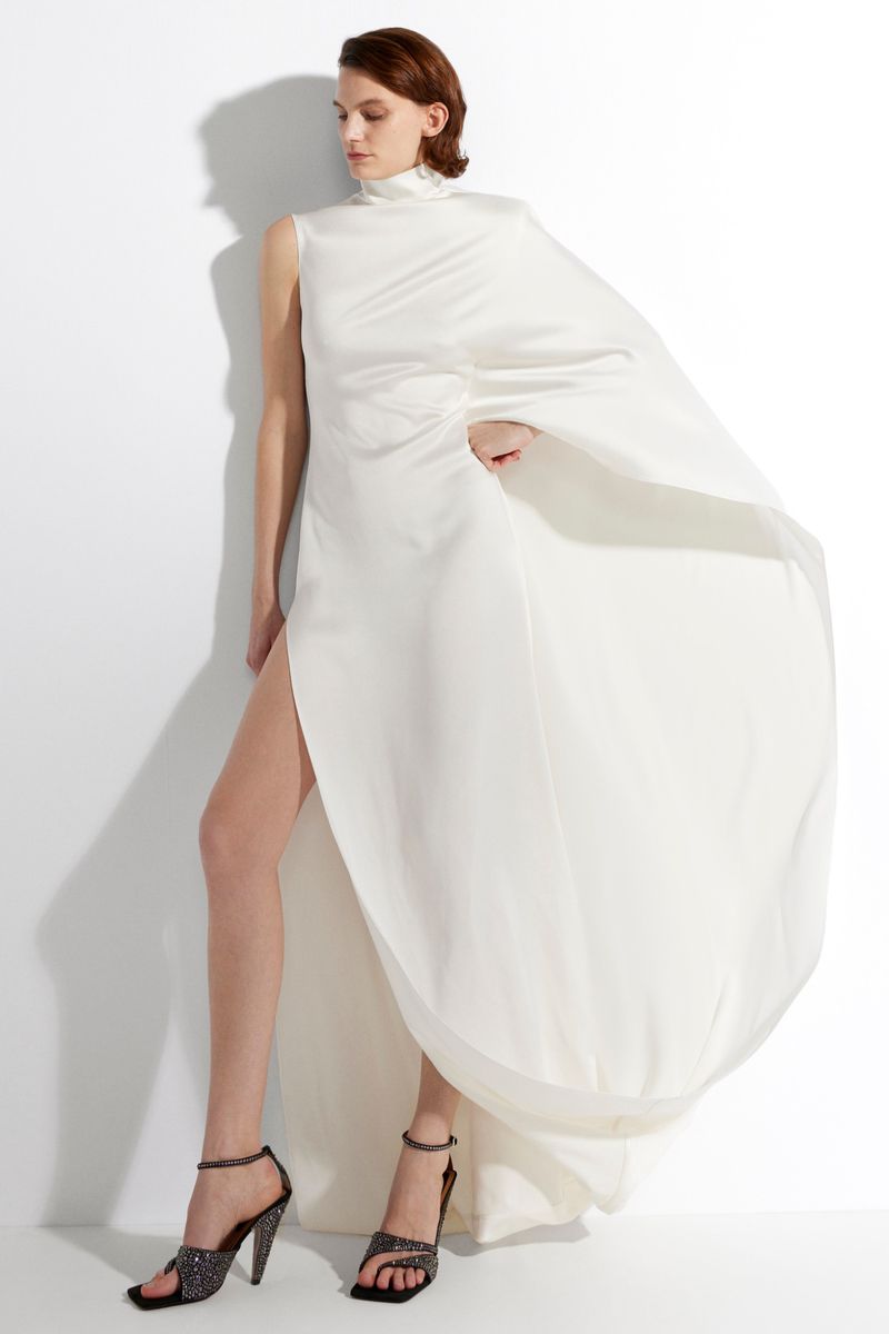 Elegant adult female posing in a floor-length, ivory gown with high-slit detail and embellished heeled sandals against a white background.
