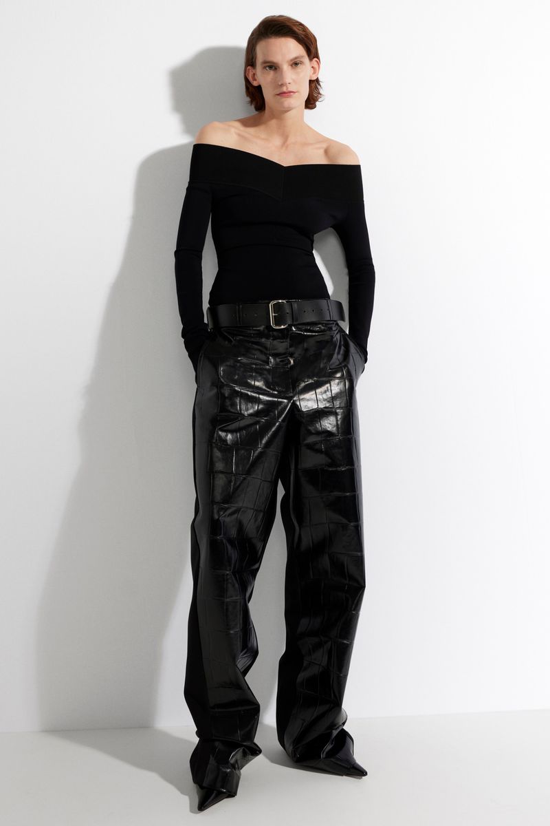 A slender woman is shown in a studio shot wearing a black off-the-shoulder top and black textured pants, standing against a white backdrop.