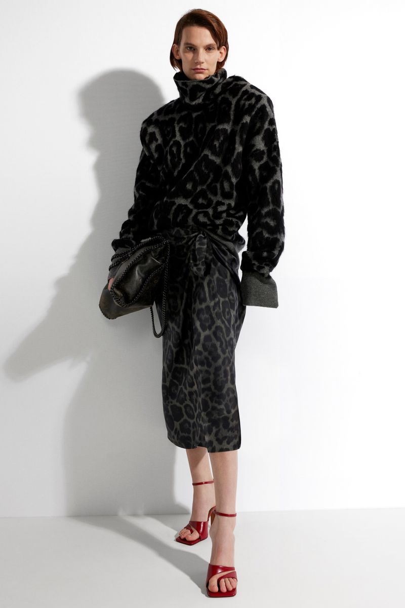 Fashionable adult female poses in a grey and black leopard-print sweater and skirt with red heels in a stark white studio.