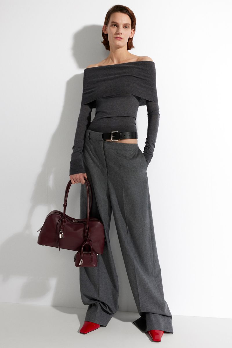 Fashion shot of a woman in a gray off-the-shoulder top and wide-leg pants, holding a burgundy satchel bag with matching miniature bag.