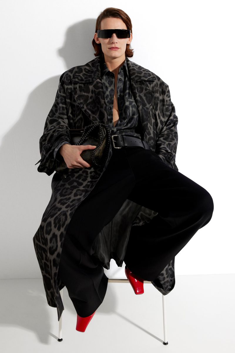 An adult model sits in a studio wearing a spotted long coat, open shirt, wide-legged pants, red high-heeled shoes, and sunglasses, holding a black purse.