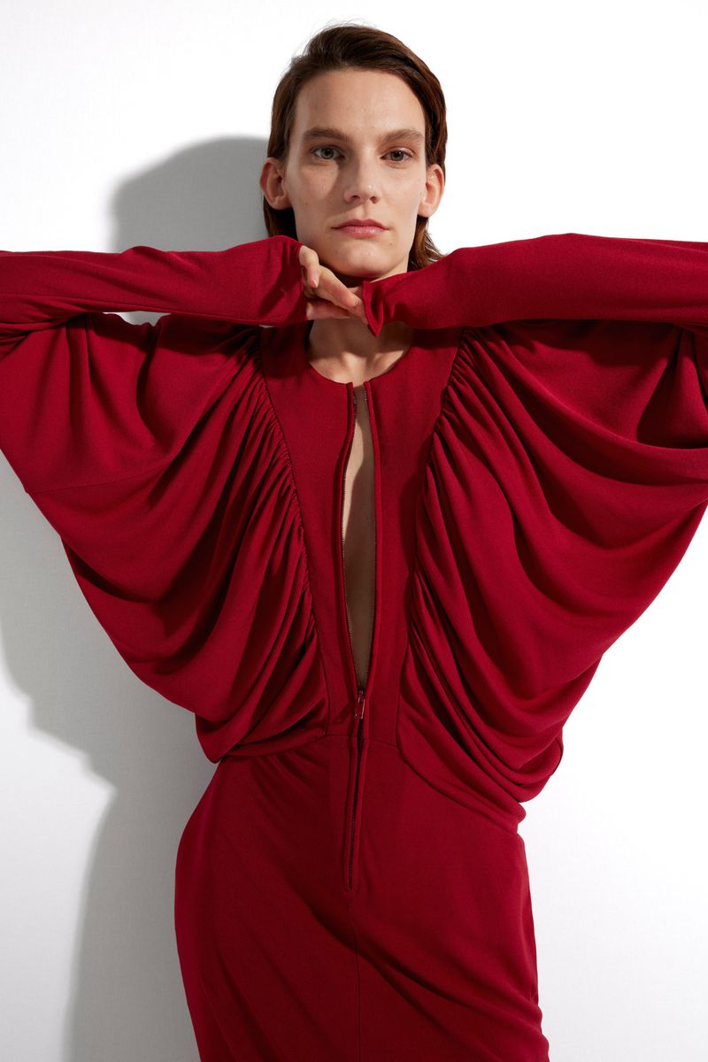 An adult female poses in a studio wearing a long sleeve, red dress with gathered fabric and a zipper front against a white backdrop.