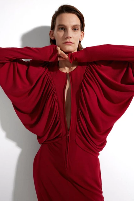 An adult female poses in a studio wearing a long sleeve, red dress with gathered fabric and a zipper front against a white backdrop.