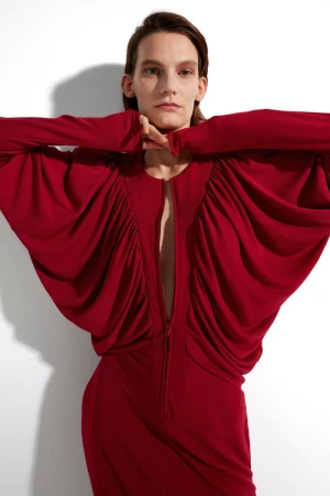 An adult female poses in a studio wearing a long sleeve, red dress with gathered fabric and a zipper front against a white backdrop.
