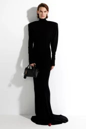 Model in a black turtleneck maxi dress holding a black handbag against a white studio wall