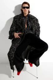 Model seated on a white chair wearing a grey leopard print coat, black trousers, sunglasses, holding a black handbag.