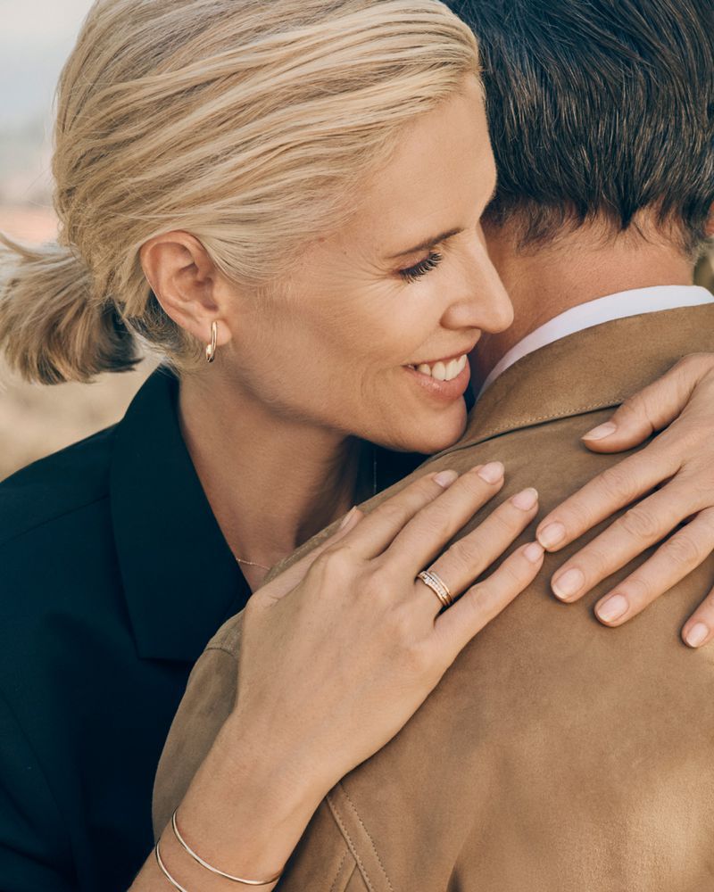 Smiling mature woman in a dark jacket hugs a man in a tan jacket outdoors, showcasing rings and bracelets in a lifestyle fashion scene.