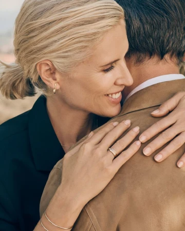 Model Judit Masco smiling as she embraces a man in a camel jacket, showcasing delicate gold earrings, a ring, and bracelets.
