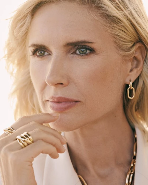 Close-up of a fair-skinned woman with blonde hair, adorned with gold jewelry, gazing off-camera against a bright, neutral background.