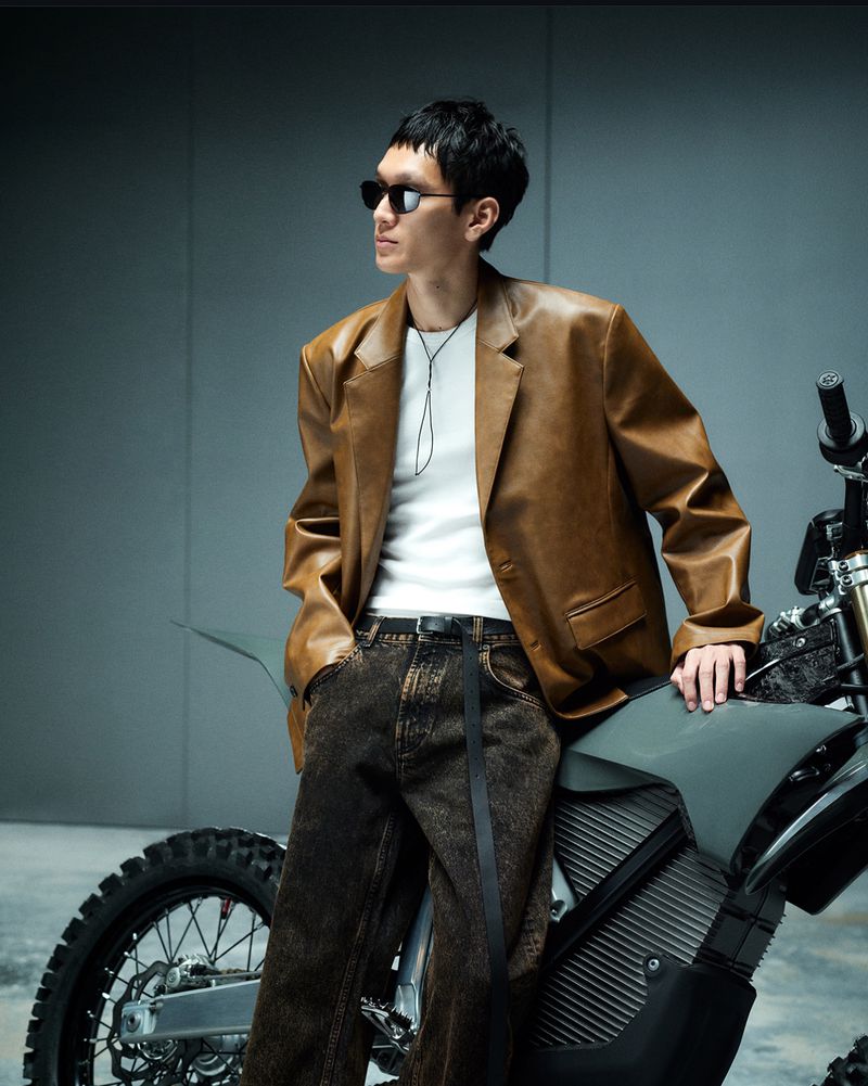 Adult male model in a brown blazer and jeans leans against a gray motorbike inside a studio with a plain backdrop and soft lighting.