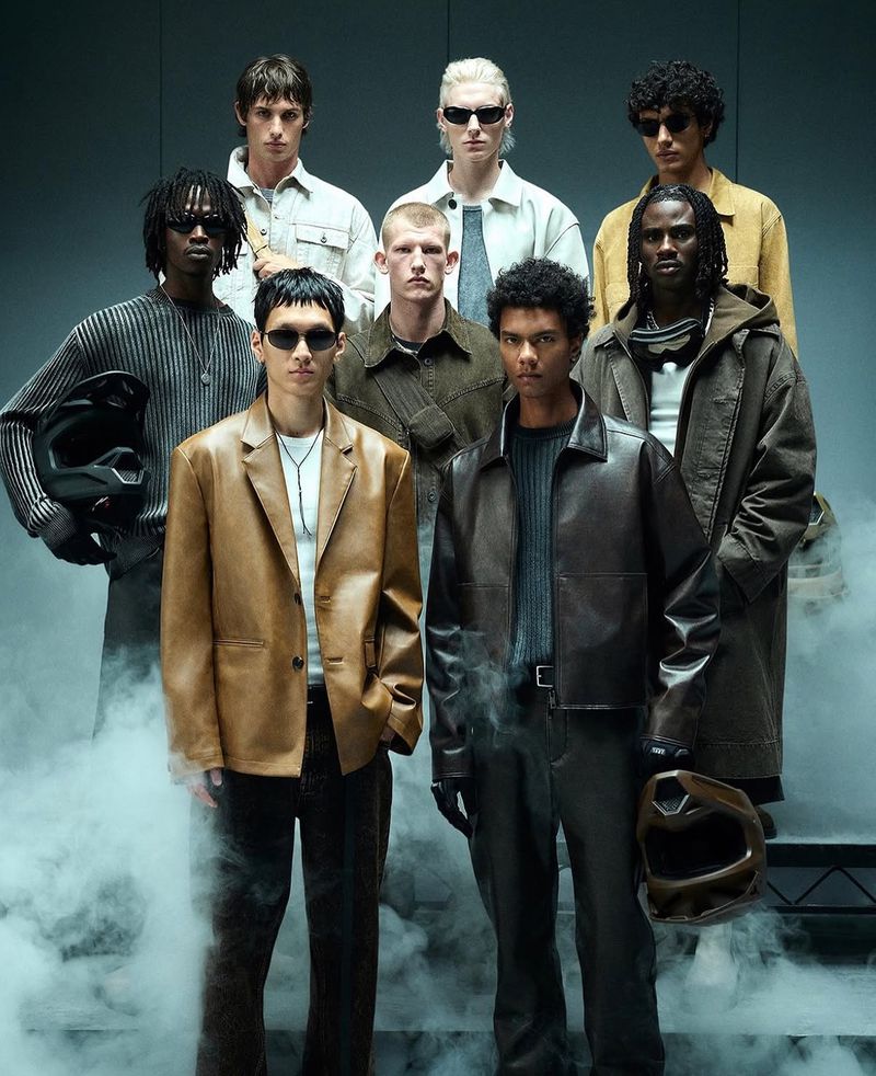 A group of men wearing different jackets and outerwear are gathered in a studio setting with diffused lighting and smoky atmosphere.
