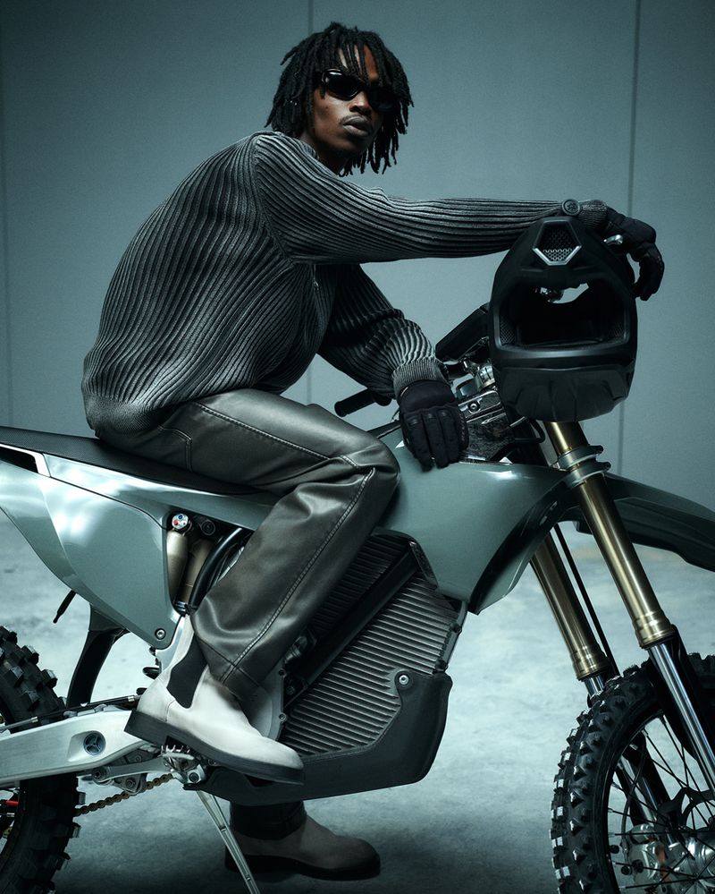 An adult Black male model poses on a silver dirt bike in a studio, wearing sunglasses, a grey sweater, leather pants, and white Chelsea boots.