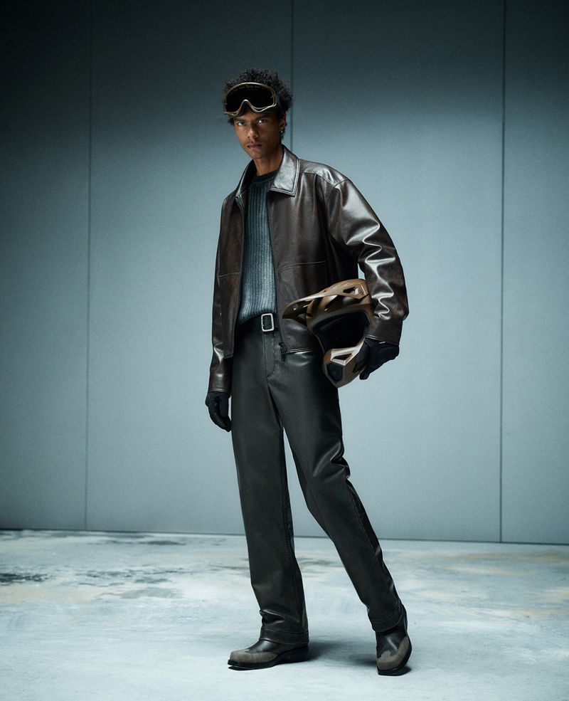 Full-length studio shot of an adult male wearing a brown leather jacket, pants, boots, and goggles, holding a motorcycle helmet.