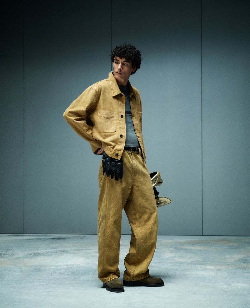 A young adult male with curly hair poses in a studio wearing a brown jacket, pants, and black gloves with metallic gold shoes.