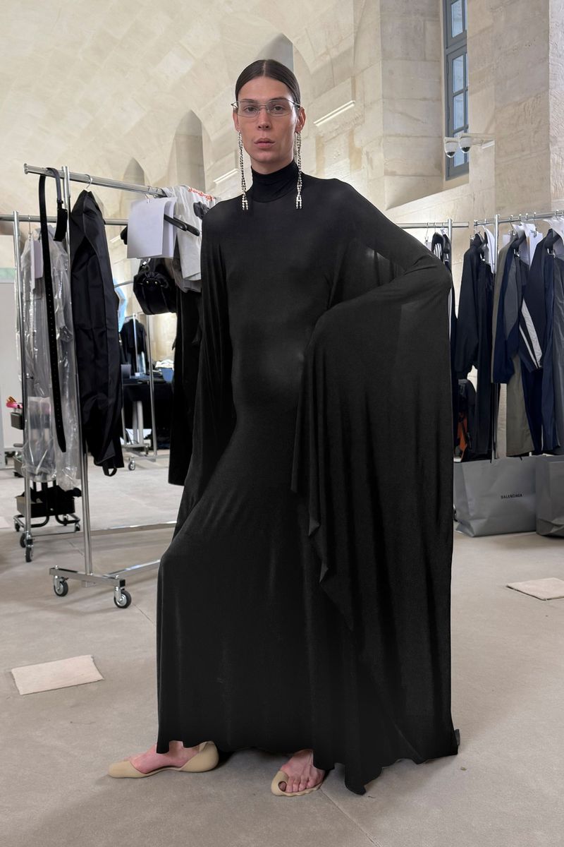 Model in black gown, wearing glasses and long earrings, posing in front of clothing racks in a studio with high ceilings and light walls.