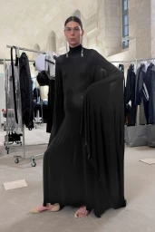 Model wearing a long black high-neck dress and silver earrings standing in a backstage room with clothing racks.