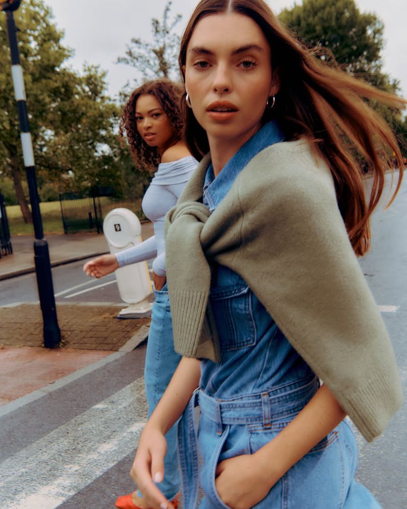 Two fashionable women walk on a crosswalk in denim and light blue clothing, with greenery and trees in the background.