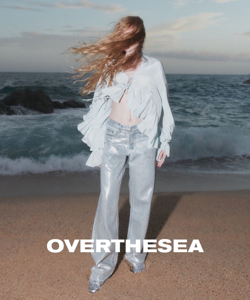 Fashion shoot on a beach with a fair-skinned adult woman wearing a light blouse and shiny pants, wind blowing her long red hair.