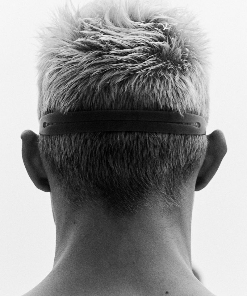 Black and white studio shot from behind of fair-skinned adult with blond hair and a black rubber head strap.