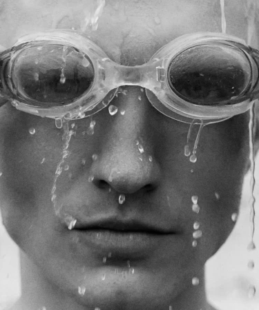 Close-up in grayscale of a person wearing swimming goggles, with water droplets streaming down their face and head.