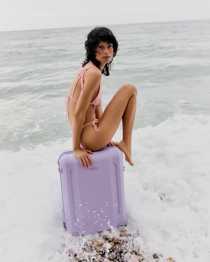 A brunette woman in a pink bikini sits on a light purple suitcase in ocean waves. The scene is set on a cloudy day at the beach.