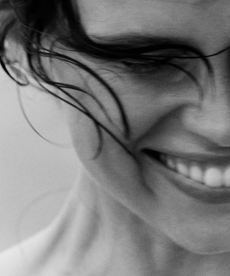 Close-up black and white portrait of a smiling adult female with dark, wet hair and white teeth against a neutral background.