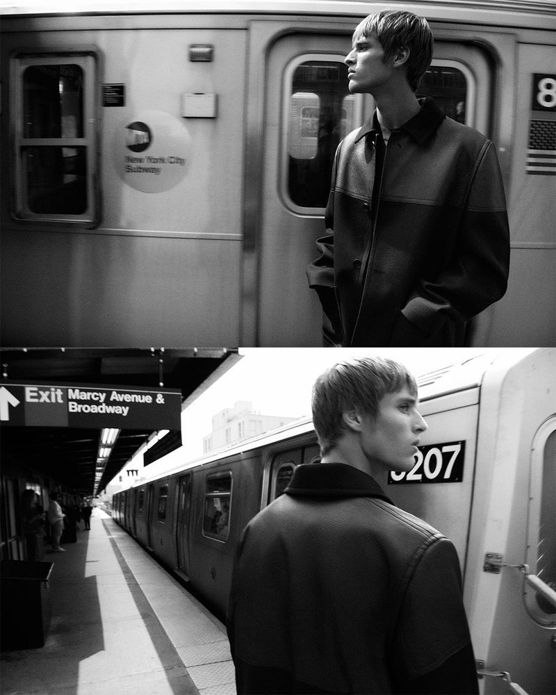 Monochrome fashion shot of a young man in a jacket standing at a subway station platform next to a subway train in profile view.