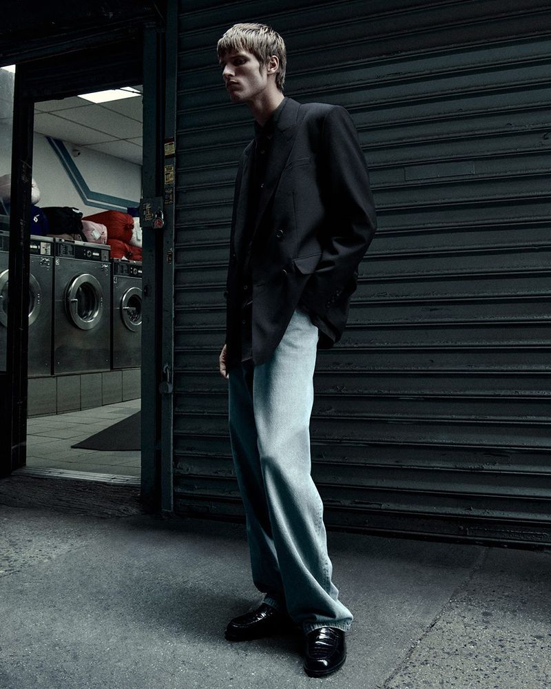 Male fashion model in a black suit and light blue jeans standing outside a laundromat with washing machines in the background.