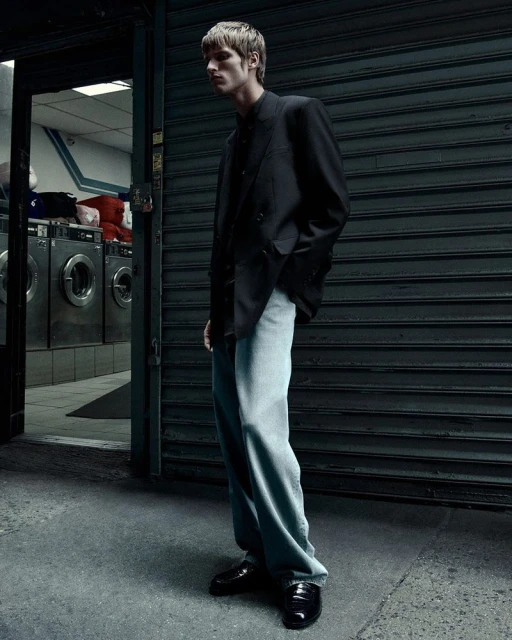 Male fashion model in a black suit and light blue jeans standing outside a laundromat with washing machines in the background.