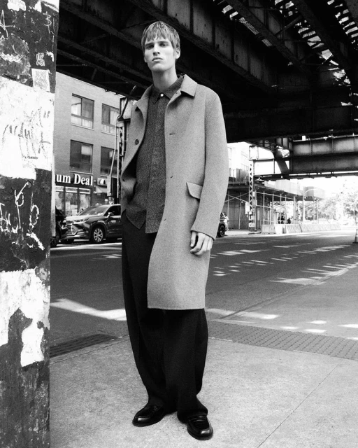 Monochrome full shot of an adult male in an overcoat standing on a city sidewalk beneath an elevated train structure on a sunny day.