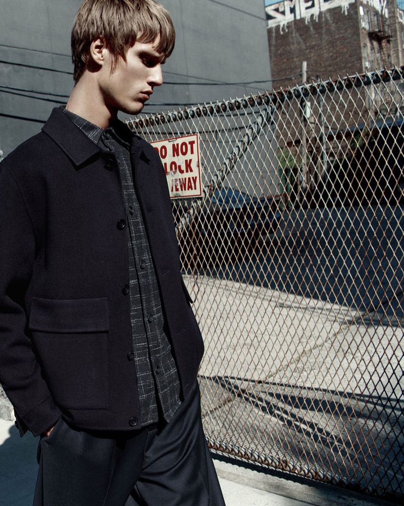 A fair-skinned adult male stands in front of a chain-link fence wearing a dark jacket and button-down shirt in an urban setting.