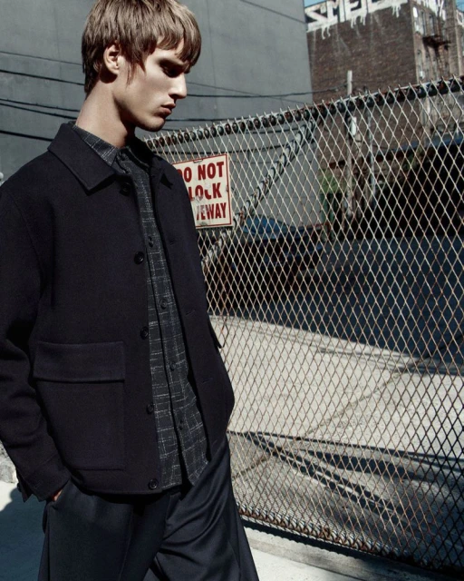 A fair-skinned adult male stands in front of a chain-link fence wearing a dark jacket and button-down shirt in an urban setting.