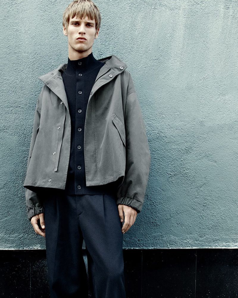Man in a grey hooded jacket and dark pants standing against a textured blue-grey wall, minimalist fashion editorial shot with soft lighting.