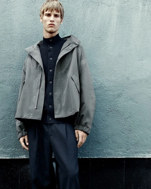 Man in a grey hooded jacket and dark pants standing against a textured blue-grey wall, minimalist fashion editorial shot with soft lighting.