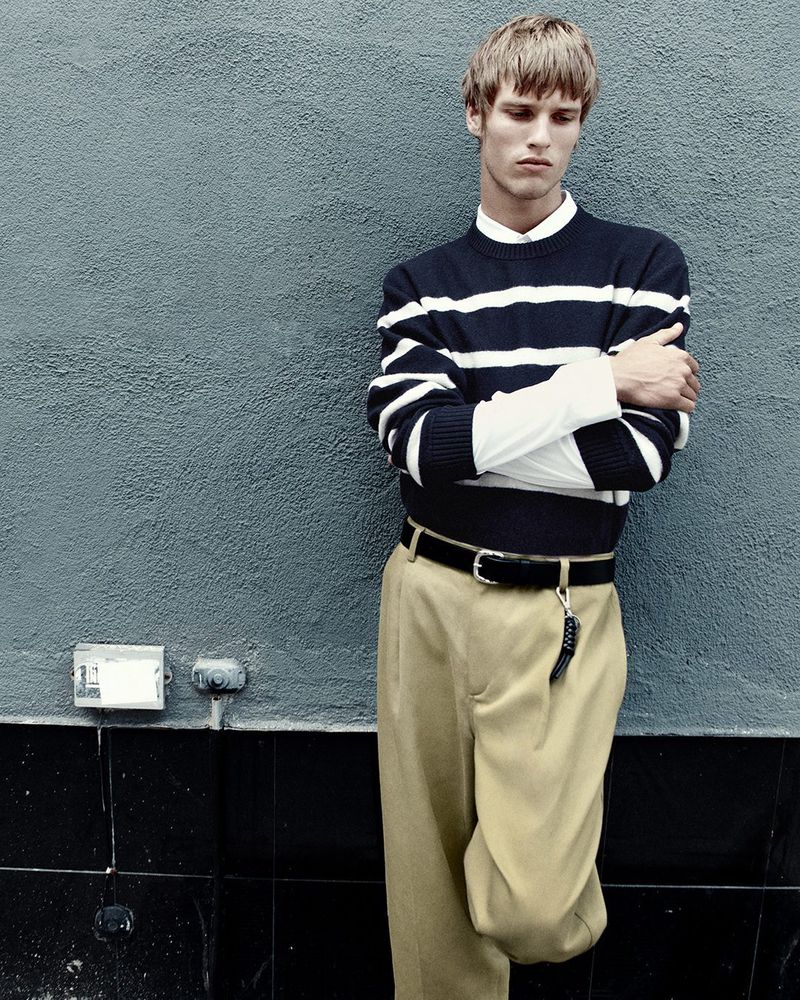 Blonde-haired male fashion model in a striped sweater, khaki pants, and black belt stands against a gray textured wall in an outdoor setting.