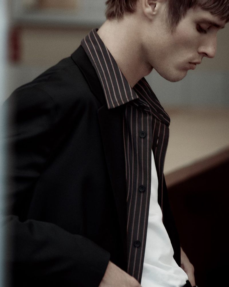Portrait of an adult male wearing a striped collared shirt and a black suit jacket in an indoor environment with neutral lighting.